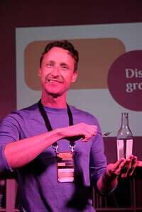 Conference speaker smiling on stage holding a glass bottle and microphone at Coffeehouse Conference.
