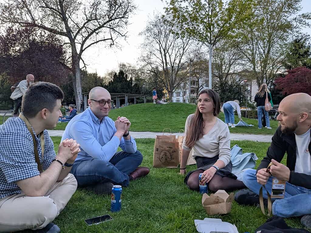 Group of diverse people sitting on grass, engaging in discussion outdoors during Coffeehouse Confere.